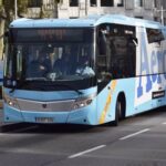 A blue and white Aerobus stopped in Plaza España, Barcelona, with passengers around, in front of the majestic fountain and with the mountain of Montjuïc in the background.