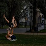 Two people in sportswear take advantage of the green spaces of the park through physical exercise. They are shown performing a yoga pose where one person holds the other with their feet while the other is lifted upside down. Learn how to describe the purpose of the image (opens in a new tab). Leave it empty if the image is purely decorative.