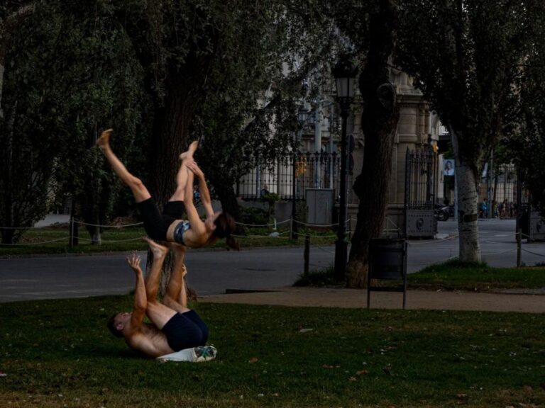 Two people in sportswear take advantage of the green spaces of the park through physical exercise. They are shown performing a yoga pose where one person holds the other with their feet while the other is lifted upside down. Learn how to describe the purpose of the image (opens in a new tab). Leave it empty if the image is purely decorative.