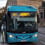 A blue and white Aerobus parked in Plaza Catalunya, with passengers boarding and alighting, in the center of Barcelona, standing out as an important connection point between the airport and the city.