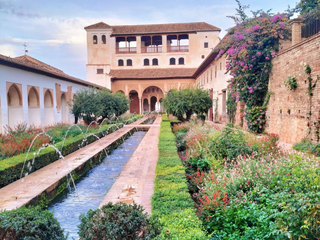 Image in one of the emblematic gardens of the Alhambra where you can observe the construction of a house, a fountain walkway, and vegetation surrounding it.
