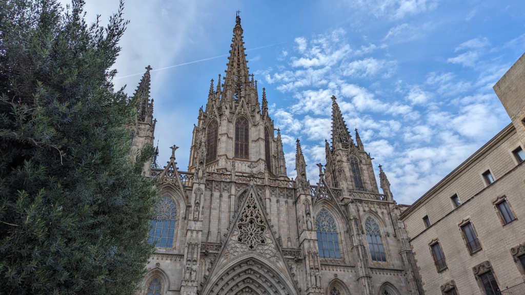 Gothic-style façade of the Barcelona Cathedral with towering spires and detailed stone carvings, set against a bright blue sky with scattered clouds and framed by green foliage on the left.