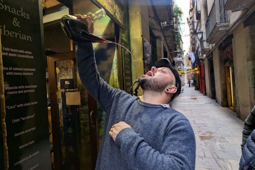 A man in a narrow street pours wine from a bottle directly into his mouth, demonstrating a traditional Catalan drinking method.
