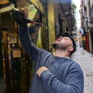 A man in a narrow street pours wine from a bottle directly into his mouth, demonstrating a traditional Catalan drinking method.